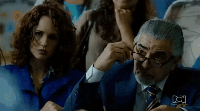 a man in a suit and tie adjusts his glasses in front of a woman in a blue shirt