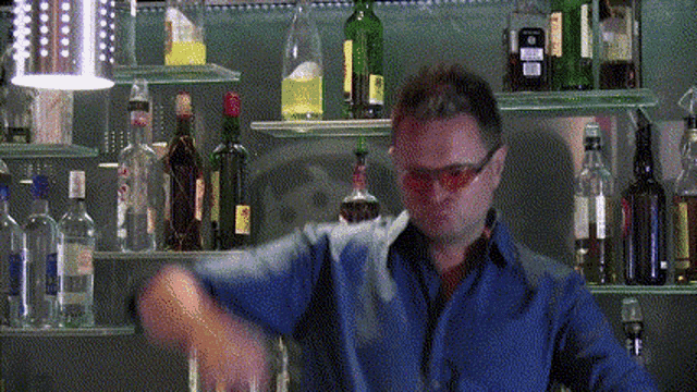 a man in a blue shirt is shaking a glass in front of a shelf full of bottles