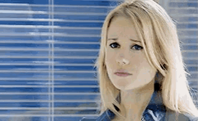 a woman with blonde hair is standing in front of a window with blinds and looking at the camera .