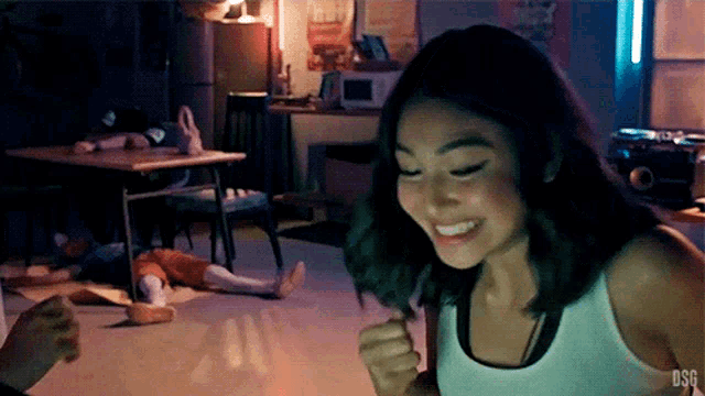 a woman in a white tank top is smiling in a dark room with a doll on the floor .