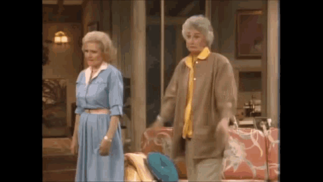 two older women are standing next to each other in a living room in front of a couch .