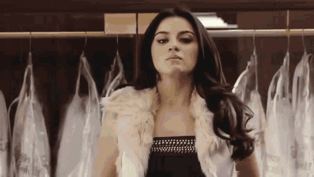 a woman wearing a fur vest stands in front of a rack of dresses