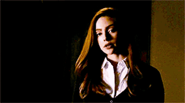 a woman with long red hair is standing in a dark room wearing a black jacket and a white shirt .