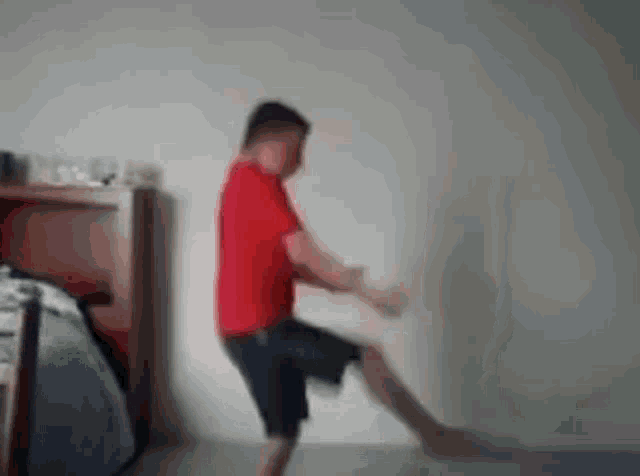 a man in a red shirt and shorts is standing on one leg in a room .