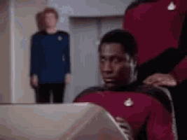 a man in a red uniform is sitting in front of a computer while another man stands behind him .