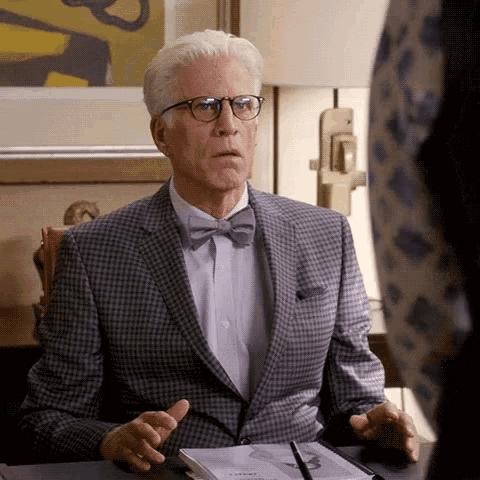 a man in a suit and bow tie is sitting at a desk talking to someone .