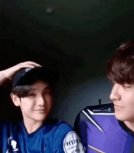 two young men are standing next to each other in a dark room . one of the men is wearing a baseball cap .