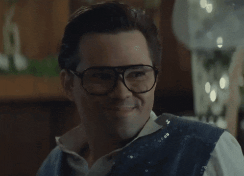 a man wearing glasses and a sequined vest smiles at the camera