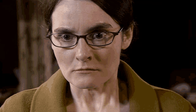 a woman wearing glasses and a brown coat is looking at the camera