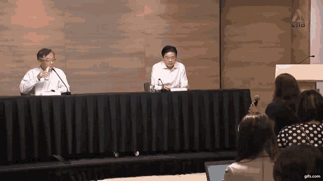 two men are sitting at a table with microphones in front of a podium with a gifs.com logo in the corner