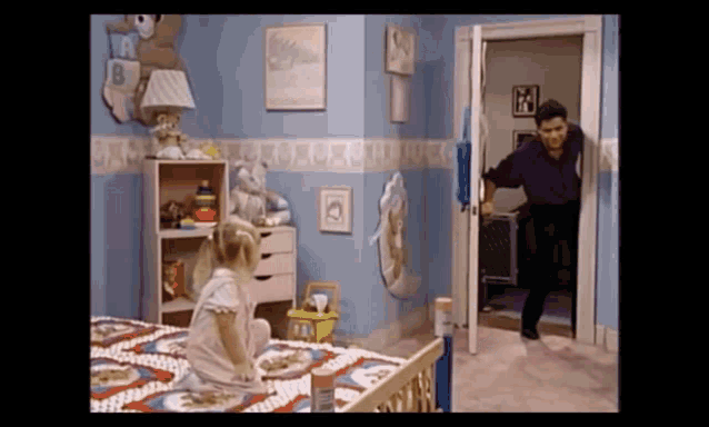 a little girl is sitting on a bed in a room with a man walking in