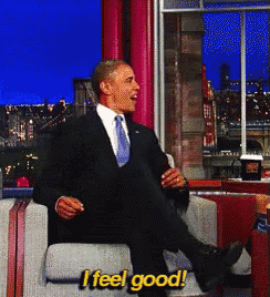 a man in a suit and tie sits in a chair with his legs crossed and says " i feel good "