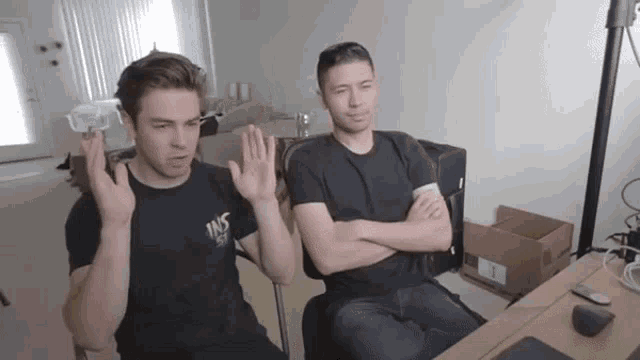 two men are sitting at a table with their arms crossed and one has a shirt that says ins on it
