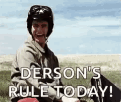 a man wearing a helmet and sunglasses is sitting in a field with the words `` derson 's rule today '' .