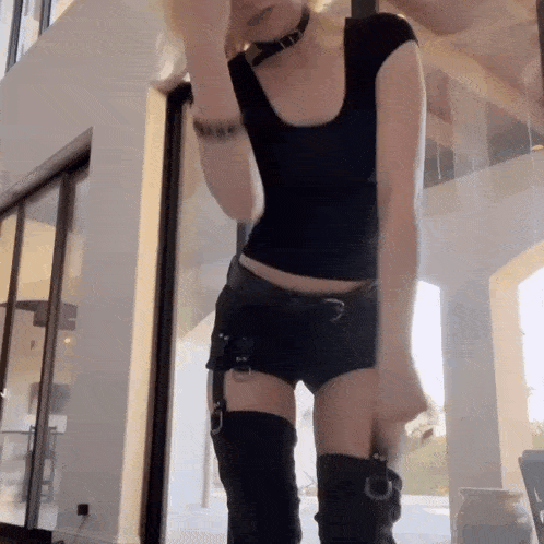 a woman wearing a choker , shorts , thigh high boots and a black top is dancing in a room .