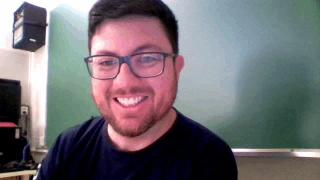 a man wearing glasses and a black shirt is smiling in front of a green board