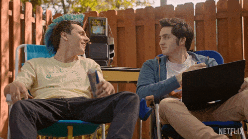 a man with a blue mohawk sits in a chair next to another man with a laptop that says netflix