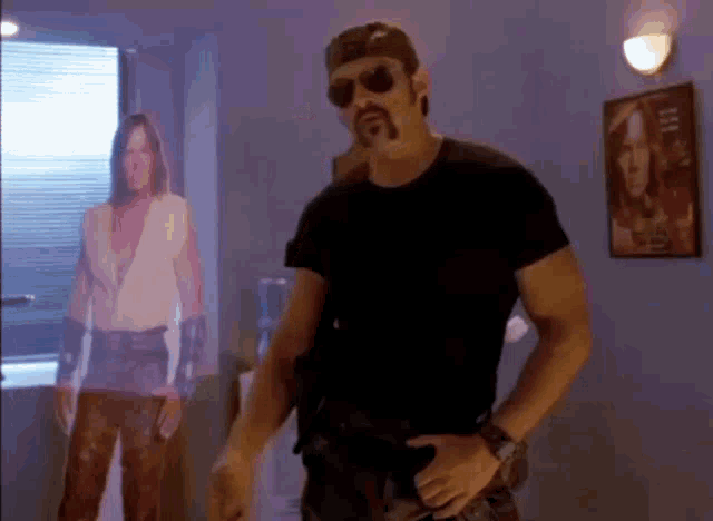 a man with a mustache and sunglasses is standing in a room with a picture of a man behind him .