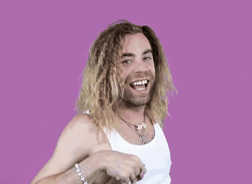 a man with long hair and a beard is wearing a white tank top and pointing at the camera .