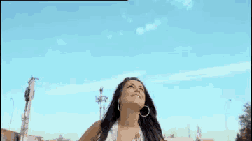a woman in a white tank top and hoop earrings smiles in front of a blue sky