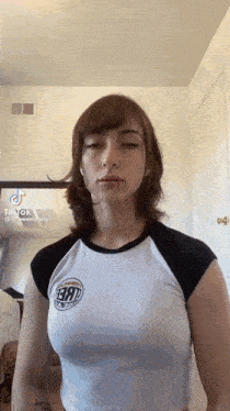 a woman in a white and black shirt is standing in front of a mirror and looking at the camera .