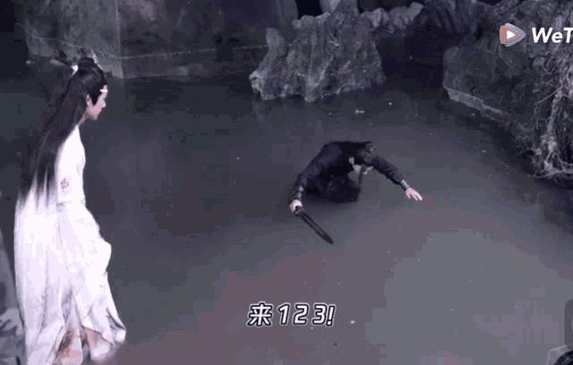 a man is kneeling in the water holding a sword while another man looks on .