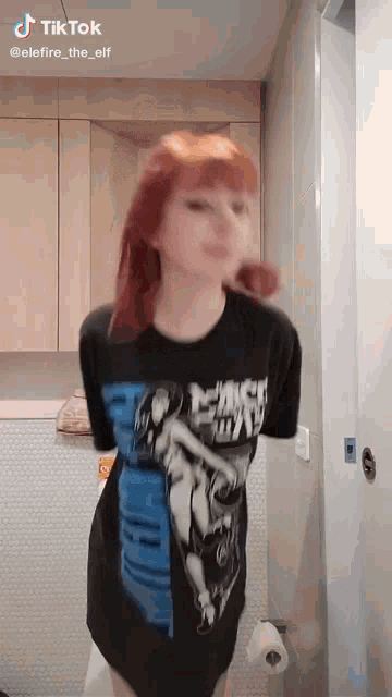 a woman with red hair is standing in a bathroom wearing a black t-shirt .