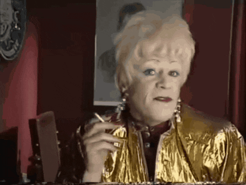 a woman in a gold jacket smoking a cigarette in front of a mirror
