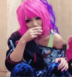 a woman with pink and purple hair is sitting down and covering her mouth with her hands .