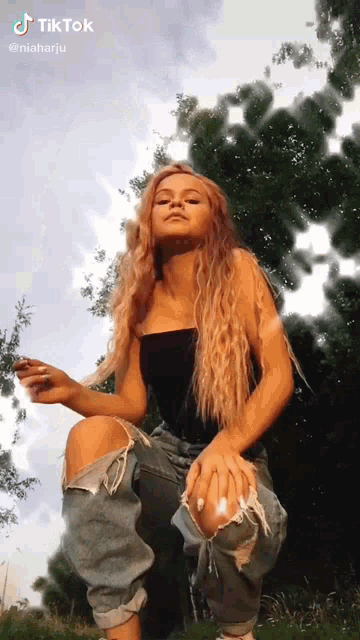 a woman in ripped jeans and a black top is sitting in the grass with trees in the background .