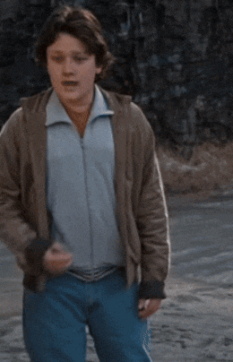 a young boy wearing a brown jacket and blue jeans stands on a dirt road
