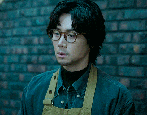 a man wearing glasses and an apron stands in front of a black brick wall
