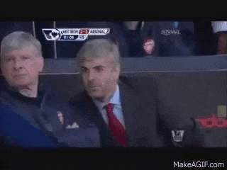two men are sitting in a stadium watching a soccer game and one of them is wearing a suit and tie .