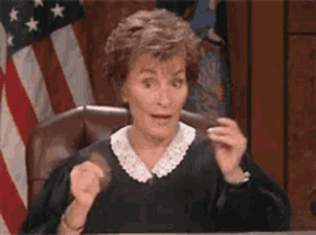 a judge is sitting at a desk in front of an american flag and making a funny face .