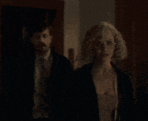 a woman wearing glasses and a black jacket stands in front of a man in a suit