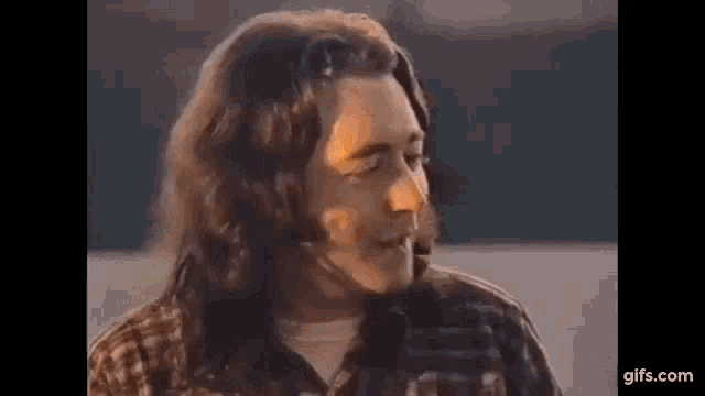 a man with long hair is standing next to a body of water and smiling .