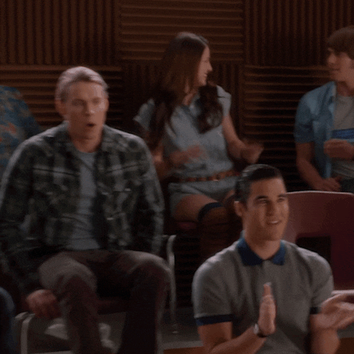 a man in a grey polo shirt is sitting in a room with other people