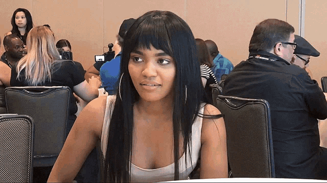 a woman in a white tank top sits at a table in a room with other people