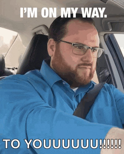 a man wearing glasses and a blue shirt is driving a car and says i 'm on my way to you