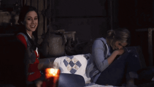 two women sit on a couch in a dark room with a candle in the foreground