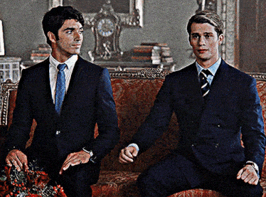 two men in suits are sitting on a couch and holding hands
