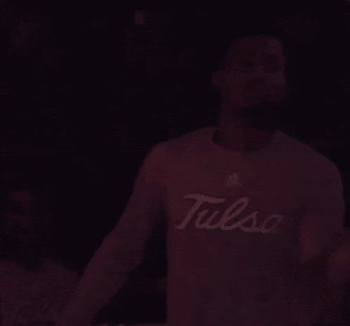 a man wearing a tulsa shirt is standing in a dark room