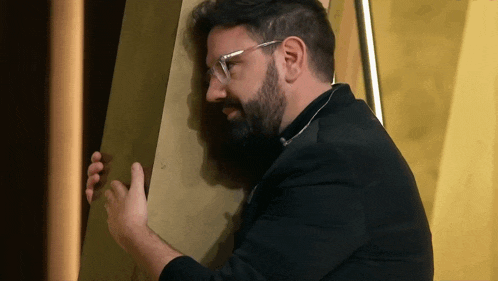 a man with a beard and glasses leans against a wall