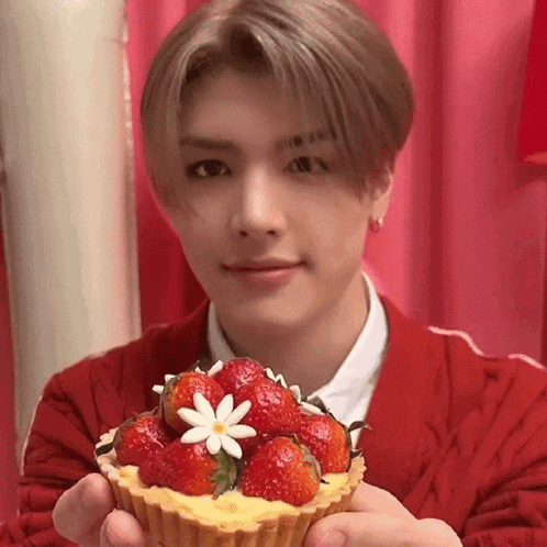 a young man in a red sweater is holding a cupcake with strawberries and a flower on top .