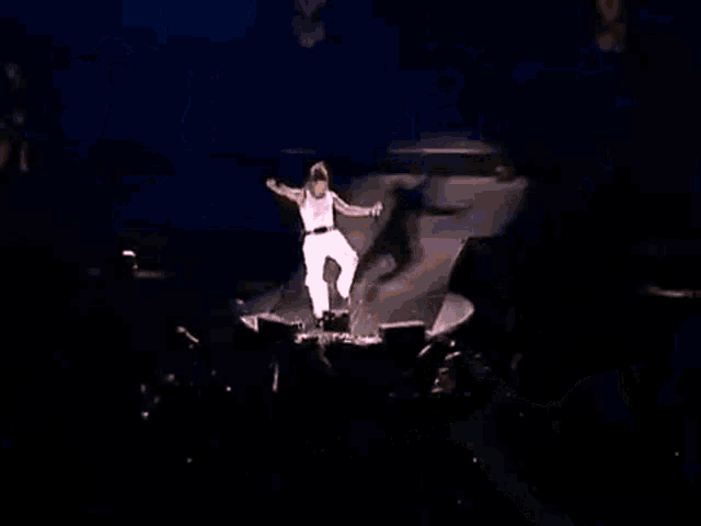 a man in a white tank top and white skirt is dancing on a stage with his arms outstretched .