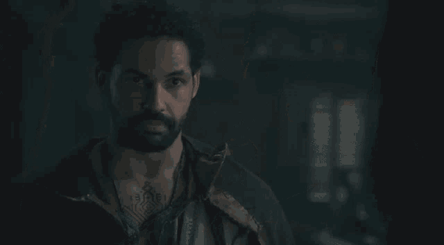 a man with a beard is wearing a leather jacket and looking at the camera in a dark room .