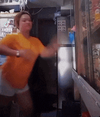 a woman in an orange shirt is dancing in front of a refrigerator in a kitchen .