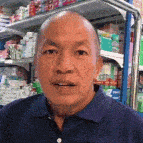 a bald man in a blue shirt is standing in front of a store shelf with boxes of toilet paper on it .