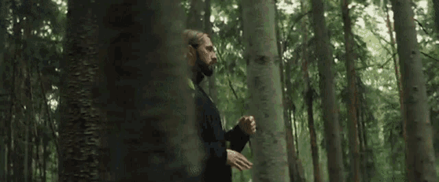 a man with long hair is standing in the middle of a forest .
