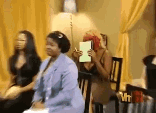 a group of women are sitting in chairs and one woman is holding a piece of paper over her face .